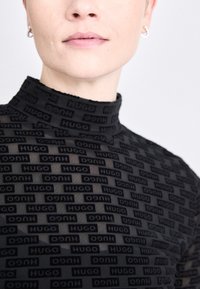 Black mesh turtleneck top featuring an all-over repeating "HUGO" logo design. The texture is sheer with a fitted silhouette and rounded neckline.