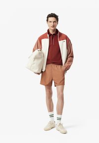 Sportig outfit för män med en färgblockerad jacka i kräm och rost, en vinröd polo, bruna shorts och vita skor med gröna accenter.