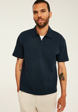Mand iført en mørkeblå kortærmet poloshirt og beige bukser, der viser en farverig øjetatovering på sin venstre underarm.