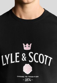 Camiseta de algodón negra con un logo blanco que presenta gráficos de una corona rosa y una rosa. Diseño de cuello redondo, textura acanalada en el collar.