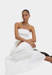Completo bianco a due pezzi composto da un top smock senza spalline e una lunga gonna con dettagli in pizzo e pieghe. Modella seduta, sandali color pelle.