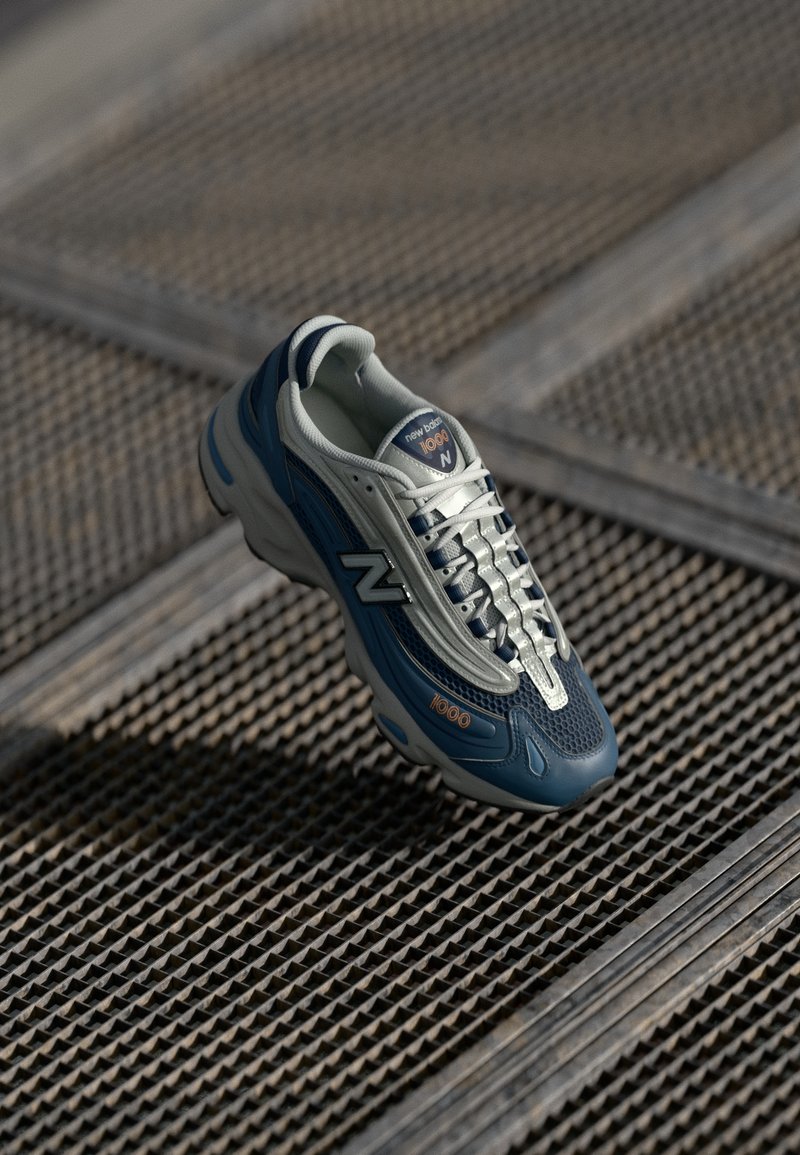 Kék és szürke New Balance 1000 sportcipő, hálós felsőrésszel, texturált talppal, fényvisszaverő részletekkel és hangsúlyos fűzős rendszerrel.