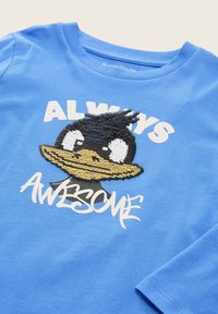Lichtblauw langemouwen t-shirt met een zwart en geel eend graphic en witte tekst met de tekst "ALTIJD GEWELDIG" in een speelse lettertype.