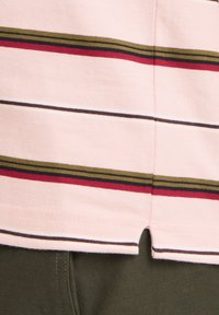 Gros plan sur un tissu rose avec des rayures horizontales vertes, rouges et noires au-dessus d'un tissu texturé vert foncé, montrant une petite fente latérale à l'ourlet.