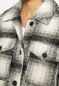 Donna che indossa un cappotto di lana a scacchi beige, nero e bianco con grandi bottoni neri e tasche a falda anteriori su una maglietta bianca.