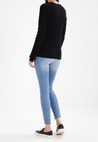 Svart texturerad tröja med långa ärmar, matchad med ljusblå skinny jeans och svarta slip-on skor, sedd bakifrån.