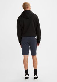 Hanorac negru cu glugă, cu o croială lejeră, asortat cu pantaloni scurți bleumarin. Modelul poartă șosete negre și adidași negri/albi, văzut din spate.