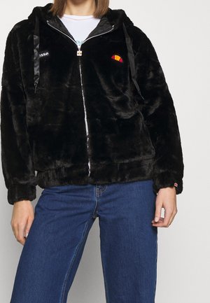 Personne portant une veste noire en peluche à capuche zippée avec les logos de la marque, une chemise blanche et un jean bleu, debout devant un fond uni.