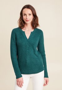 Maglione a coste in teal con maniche lunghe, scollo a V e patta con cinque bottoni. Presenta motivi testurizzati e una leggera lucentezza. Indossato con pantaloni bianchi.