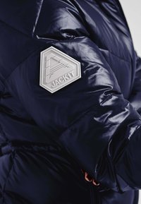 Marineblå pufferjakke med en glat tekstur har et fremtrædende hvidt sekskantet mærke mærket "JACK1T" på venstre ærme.