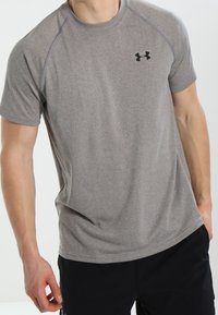 Muž v šedém tričku s krátkým rukávem sportovního střihu s černým logem Under Armour a v černých šortkách, stojící na neutrálním pozadí.