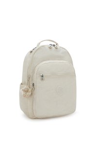 Mochila de tela beige con un diseño redondeado, bolsillo frontal con cremallera y un llavero de mono de peluche decorativo adjunto. Presenta el logotipo en la parte frontal.