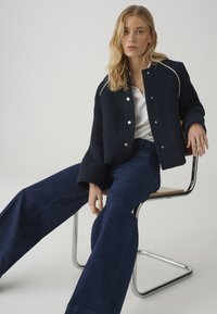 Woman with blonde hair sitting on a metal and wood chair, wearing a navy blue jacket, white top, and wide-leg dark blue jeans.