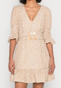 Donna che indossa un vestito beige a fiori con scollatura a V, maniche a palloncino, orlo arricciato e fibbia quadrata in vita.