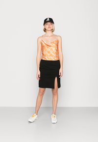 Top de gola de cowl com padrão laranja combinado com uma saia lápis preta com uma fenda lateral. O modelo usa ténis brancos e um boné preto.