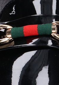 Correa de material negro brillante con herrajes dorados, destacada por una banda tejida verde y roja en el centro. Textura suave con un ligero brillo.