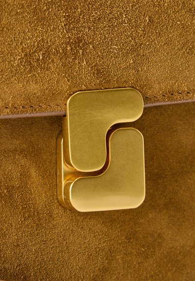 Κοντινή λήψη ενός κούμπωμα με χρυσή απόχρωση σε καφέ σουέντ υλικό, που δείχνει την υφή και το ράψιμο κατά μήκος της άκρης.