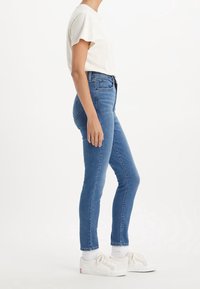 Jeans de mezclilla azul de cintura alta y corte ajustado, con cinco bolsillos y dobladillos enrollados, combinados con una camiseta de color claro y zapatillas blancas.