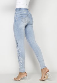 Femme portant un jean skinny bleu clair brodé et des sandales à talons hauts transparentes, debout devant un fond uni.