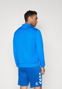 Blå atletisk jakke med høj krave og tekstureret stof. Matcher med blå shorts med hvide chevron-striber og logo.