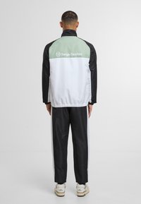 Träningsjacka i svart, vitt och mintgrönt, med en logotyp på baksidan. Slät tyg med dragkedja i kragen och elastisk midja.