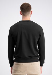 Felpa nera con maniche lunghe, colletto e polsini a coste, con un piccolo dettaglio di texture liscia sul retro. Abbinata a pantaloni chiari.