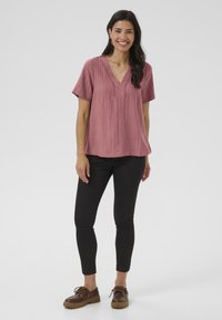 Haut rose à manches courtes avec un col en V et un tissu texturé, associé à un pantalon noir ajusté et des chaussures à lacets marron.
