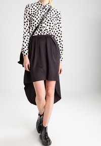 Weiße Bluse mit schwarzem Herzmuster, schwarzer plissierter asymmetrischer Rock und schwarze Lackleder-Stiefeletten, kombiniert mit einer schwarzen Tasche.