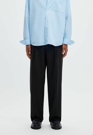 Personne portant une chemise bleu clair boutonnée, un pantalon noir ample et des chaussures tissées noires, debout devant un fond blanc uni.