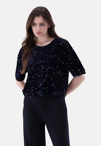 Navyblaues, mit Pailletten besetztes Oberteil mit lockerer Passform, kurzen Ärmeln und rundem Ausschnitt. Die Oberfläche ist glänzend und strukturiert mit einem durchgehenden Paillettenmuster.