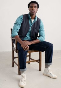 Ciemna kamizelka na tle niebiesko-zielonej w paski, długiej koszuli z długim rękawem, ciemne dżinsy z podwiniętymi mankietami i białe sneakersy typu high-top. Drewniane krzesło na jasnym tle.