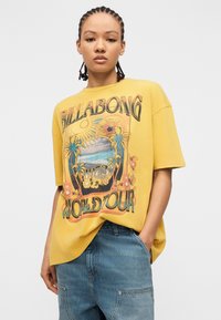 Νεαρό άτομο με κοτσίδες, φορώντας κίτρινη μπλούζα Billabong με γραφικά και μπλε τζιν, στέκεται με το ένα χέρι στην τσέπη.