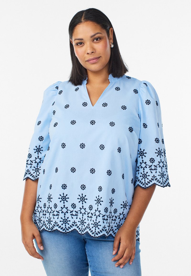 Kvinde iført en lyseblå bluse med sort blomsterbroderi og bølget kant, kombineret med mellemblå jeans, stående mod en hvid baggrund.