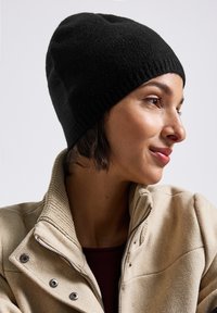Donna con un berretto di maglia nero e una giacca beige con zip, rivolta verso destra con un leggero sorriso su uno sfondo chiaro e semplice.
