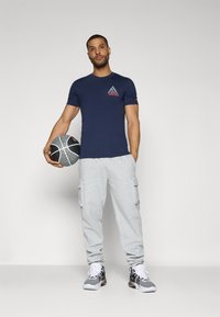 Camiseta de la marina con un logo colorido, pantalones de chándal gris con bolsillos laterales y zapatillas en blanco y negro, sosteniendo un balón de baloncesto.