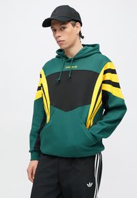 Mladý muž oblečený v zelenej, čiernej a žltej Adidas mikine s logom, čiernej Adidas čiapke a čiernych Adidas nohaviciach s bielymi pruhmi.