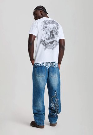 Mann mit geflochtenem Haar, der ein weißes Grafik-T-Shirt und blaue Jeans mit Schädel- und Schlangendesign trägt, steht vor einem schlichten hellen Hintergrund.