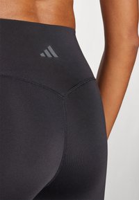 Leggings negros de talle alto con una textura suave, que presentan un detalle de costura y un discreto logo de Adidas en gris cerca de la cinturilla.