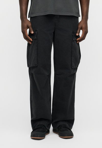 Pantalon cargo noir en coton, avec poches latérales, coupe ample et texture légèrement délavée. Associé à des baskets noires.