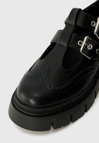 Chaussures en cuir noir avec un motif brogue, dotées de deux sangles ajustables et d'une semelle épaisse avec des rainures prononcées ; finition lisse.