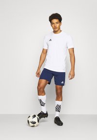 Valge lühikeste varrukatega sportlik T-särk musta Adidas logo kaubamärgiga; tumesinised lühikesed püksid; mustad jalgpalliketsid; valged sokid mustade triipudega; jalgpall.
