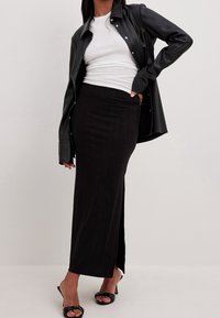 Falda maxi negra acanalada, de corte ajustado, con una abertura alta. Combinada con una camiseta blanca ajustada y una chaqueta de cuero negra tipo camisa. Textura suave.