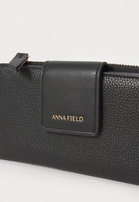 Black textured wallet with a smooth leather flap, gold "ANNA FIELD" logo, and a zip closure. Features include a practical design and accent details.
