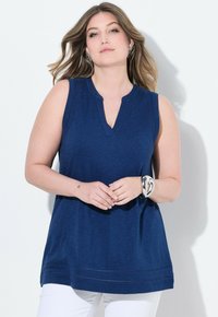 Tunic senza maniche di colore blu navy con scollatura a V e delicato dettaglio di cucitura all'orlo, realizzata in tessuto morbido. Indossata con pantaloni bianchi.