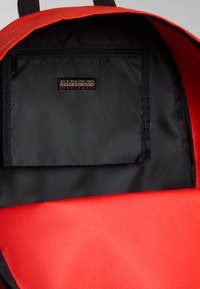 Interior de una mochila roja con un forro negro, cuenta con un bolsillo con cremallera y una etiqueta de logo. Tela duradera con un diseño estructurado.