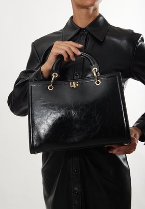 Black leather handbag with a glossy finish, featuring rounded handles, gold-tone hardware, and a logo accent on the front.