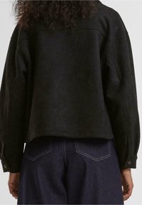 Veste en daim noir avec une coupe courte, une coupe décontractée et des manches longues. Les détails remarquables incluent des fermetures à boutons et une texture lisse.