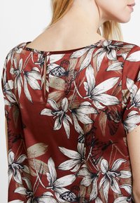 Rød floral bluse med store hvide blomster og sorte sommerfugle, med en nøglehul-rygdetalje og blødt stoftekstur.