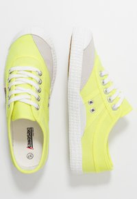 Zapatillas de lona amarillas con acentos de ante gris, suela de goma blanca, cordones planos y ojetes de metal. Tejido texturizado y diseño minimalista.