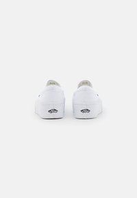 Zapatillas de lona blancas sin cordones con suela de plataforma, que presentan un logo negro en el talón y una textura suave.